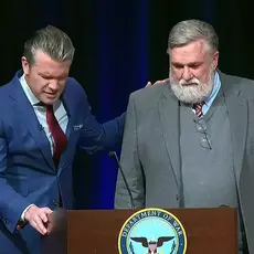 Defense Secretary Pete Hegseth and Christian nationalist pastor Doug Wilson praying at lectern bearing the logo of the "Department of War." Their heads are bowed, and Hegseth has one hand on the lectern and one on Wilson's shoulder.