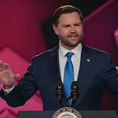 Vice President J.D. Vance gestures with his hands while speaking at lectern on stage at TPUSA's AmericaFest; the TPUSA logo is partially visible behind him.