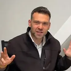 Jack Posobiec, a casually dressed white man seated at his laptop, gestures with his hands while speaking into the camera in front of a white wall background.
