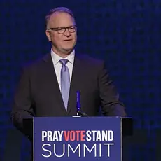 Phillip Jauregui, balding white man wearing a suit and tie, stands behind a lectern that carries the logo for the Pray Vote Stand Summit