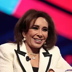 Jeanine Pirro, wearing cream colored suit with black trim, smiles while standing at lectern with red and blue stage backdrop blurred behind her.