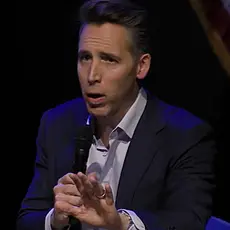 Sen Josh Hawley holds a microphone in his right had and gestures toward the audience with his left hand while seated on stage