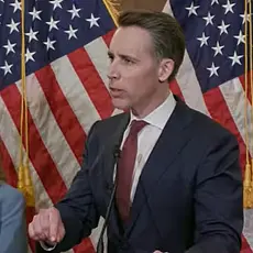 Image from press conference shows Sen. Josh Hawley in dark suit and red tie and standing in front of two U.S. flags, looking off camera at a questioner. Standing behind him on the left is Kristen Waggoner, head of the anti-abortion legal group Alliance Defending Freedom. 