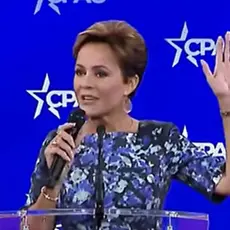 Kari Lake on stage in front of CPAC logos; she holds microphone in her right hand with her left hand raised in the air. 