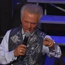 Kent Christmas, standing on stage at the Ryman Auditorium, speaks into a microphone in his right hand while pointing toward the crowd with his left hand; he looks angry.