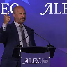Heritage Foundation President Kevin Roberts, a bald white man wearing suit and tie, gestures with his right hand while speaking behind a lectern bearing the ALEC logo, which is also repeated on the background. 