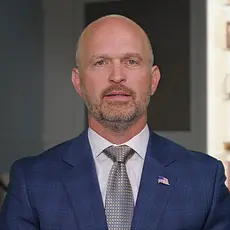 Heritage Foundation President Kevin Roberts, in suit and tie, speaks into a camera standing in what looks like an office, with a bookshelf behind him.