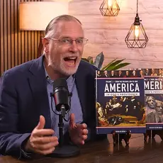 Kevin Swanson speaks animatedly into a microphone; two copies of his 1,000 page textbook on America's "Christian history" are propped up on the table next to him. 