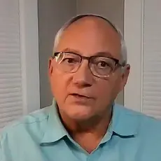 Glenn Clary, a balding white man with gray hair and wearing glasses, is seated in a nondescript room wearing a light blue polo shirt.