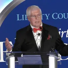 Mat Staver, a white man wearing a suit and red bowtie, gestures while speaking at lectern in front of logo for Liberty Counsel's 2025 gala and its theme "A New Birth of Freedom"