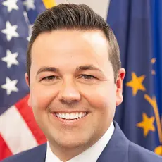 Portrait-style official photo of Indiana Lt. Gov. Micah Beckwith smiling into the camera with U.S. and Indiana flags visible behind him.