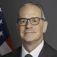 Formal portrait photo of Michael Anton in suit and tie looking into the camera with an American flag behind his right shoulder