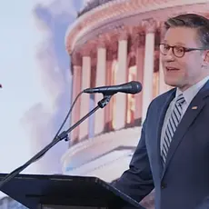 Image shows Mike Johnson standing and a lectern and speaking into a microphone in front of an illustrated image showing the Capitol dome and a U.S. flag fluttering in the breeze.