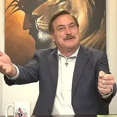 Mike Lindell, seated in front of a painting of a lion's head superimposed on the head of Christ, gestures with both hands. A cross and a US flag are visible on the shelves behind him. 
