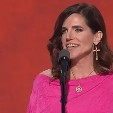 Rep. Nancy Mace, wearing bright pink dress and oversize cross-shaped earrings, speaks into a microphone in front of a red background on stage at the 2024 Republican National Convention.
