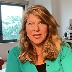 Naomi Wolf is seated in what appears to be a home office, with a window, shelf, and art visible behind her.