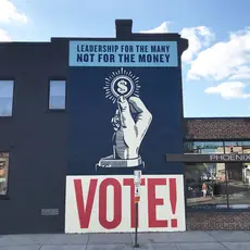 A mural with a handle holding a coin and text that reads "Leadership for the many, not for the money" and "Vote!"