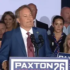 Texas Attorney General Ken Paxton stands at microphone behind a lectern with his Paxton 26 campaign logo. Supporters holding Paxton signs are gathered behind him.
