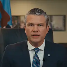 Pete Hegseth, wearing suit and tie, is seated in his office and speaking into the camera.