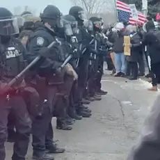 Image shows line of helmeted baton-wielding law enforcement officers facing off against protesters