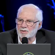 Rick Joyner preaching into a microphone while seated onstage with his laptop in front of him. s laptop.