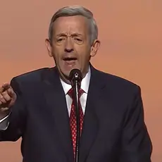 Robert Jeffress in blue suit and red tie stands at a microphone on stage at the 2026 Republican midterm convention, pointing toward the audience to emphasize a point he is making. ard the audience 