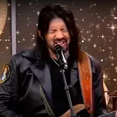 Robin Bullock, a white man with long black hair, grimace while speaking/singing into a microphone and holding a guitar. 