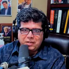 Shane Vaugh, wearing glasses and headphones, is seated in a cluttered office speaking into a microphone.