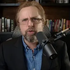 Steve Turley, a white man with red hair and beard, speaking into a microphone while seated in front of bookshelves in what appears to be a home office or library.