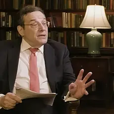 Tom Klingenstein, a white man wearing glasses and a suit and tie, holds papers in his right hand and gestures with his left. He is seated in front of bookshelves in what appears to be an office or home library. He is looking toward his left and speaking with John Eastman, who is not visible in this image cropped from video of their interview.