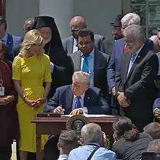 President Donald Trump seated at a desk in the White House rose garden, signing an executive order creating a Religious Liberty Commission, as White House aide Paula White and a group of religious leaders and activists look on. 