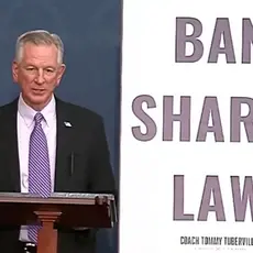 Sen. Tommy Tuberville, wearing suit and tie, stands behind lectern next to a huge poster reading BAN SHARIA LAW.