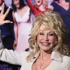 Dolly Parton, a white woman with blonde hair, wearing a white jacket and smiling and waving.