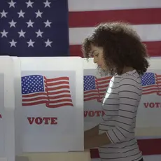 A Black woman voting at a voting booth that says "vote" on it all in front of a large American flag.