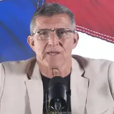 Michael Flynn speaks into a microphone seated in front of a close-up image of stars and stripes from a U.S. flag. 