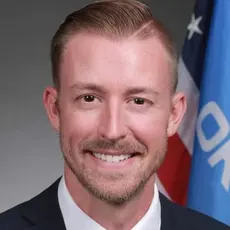 Official-style portrait of Ryan Walters smiling into the camera with US and Oklahoma flags visible over his left shoulder.
