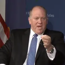 Tom Homan, in suit and tie and seated in front of an American flag, gestures while speaking. 