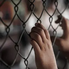 Two hands grip a chain link fence while other people are out of focus in the background.