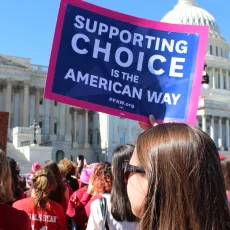 A group of people stand in front of the Capitol Building; one woman holds a sign that says "Supporting Choice is the American Way"