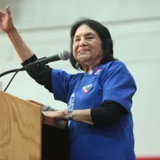 Dolores Huerta stands at a podium. She is smiling, and her right arm is lifted toward the crowd.