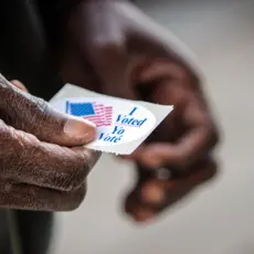A close up of an elderly Black man's hands. He has just finished casting his ballot, and is holding his “I voted” sticker.